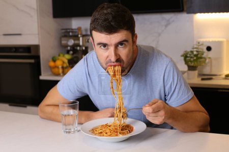 Téléchargez les photos : Portrait de beau jeune homme mangeant des spaghettis à la cuisine - en image libre de droit