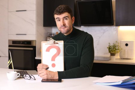 Photo for Portrait of handsome young man holding clipboard with question mark at kitchen - Royalty Free Image