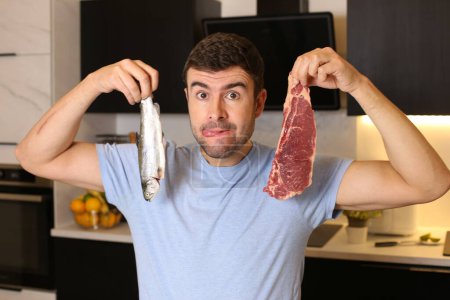 Téléchargez les photos : Portrait de beau jeune homme tenant de la viande et du poisson à la cuisine - en image libre de droit