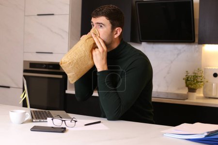 Photo for Portrait of handsome young man breathing with paper bag while working from home at kitchen - Royalty Free Image