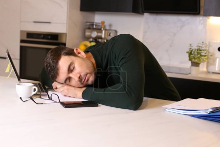 Foto de Retrato de joven guapo durmiendo en la mesa mientras trabaja desde casa en la cocina - Imagen libre de derechos
