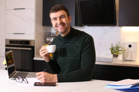 Foto de Retrato de joven guapo con taza de café trabajando desde casa en la cocina - Imagen libre de derechos