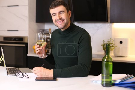 Téléchargez les photos : Portrait de beau jeune homme avec verre de vin travaillant de la maison à la cuisine - en image libre de droit