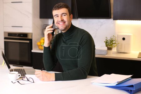 Photo for Portrait of handsome young man talking by phone while working from home at kitchen - Royalty Free Image