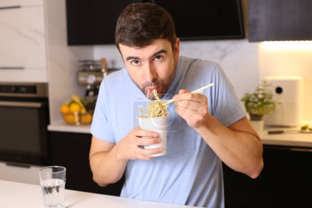 Photo for Portrait of handsome young man eating cup noodles at kitchen - Royalty Free Image