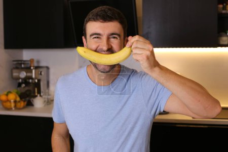 Téléchargez les photos : Portrait d'un beau jeune homme tenant une banane à la cuisine - en image libre de droit