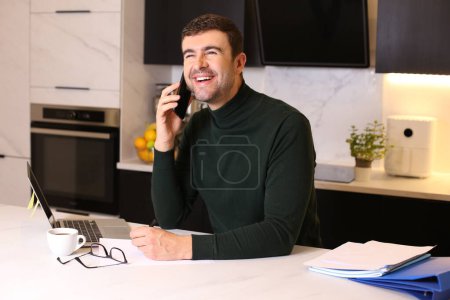 Téléchargez les photos : Portrait de beau jeune homme parlant par téléphone tout en travaillant de la maison à la cuisine - en image libre de droit