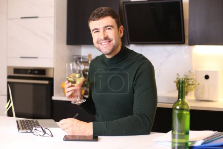 Foto de Retrato de joven guapo con copa de vino trabajando desde casa en la cocina - Imagen libre de derechos