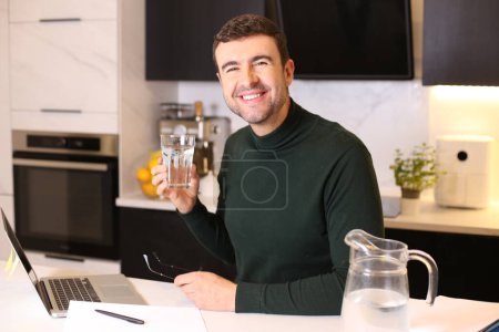 Foto de Retrato de un joven guapo bebiendo agua mientras trabaja desde casa en la cocina - Imagen libre de derechos