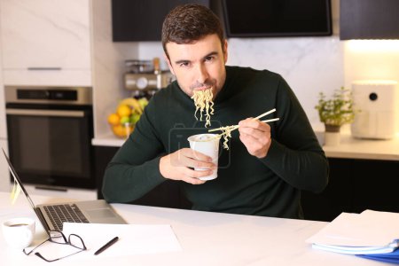 Téléchargez les photos : Portrait de beau jeune homme mangeant des nouilles de tasse tout en travaillant de la maison à la cuisine - en image libre de droit