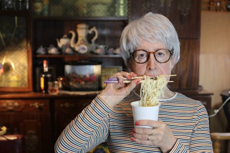 Photo for Close-up portrait of mature woman eating cup noodles at home - Royalty Free Image