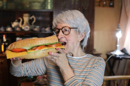 Photo for Close-up portrait of mature woman with giant sandwich at home - Royalty Free Image