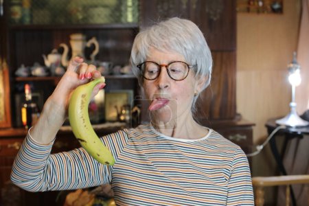 Foto de Close-up portrait of mature woman with banana sticking tongue out at home - Imagen libre de derechos
