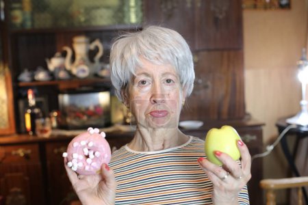 Photo for Close-up portrait of mature woman with glazed doughnut and green apple at home - Royalty Free Image