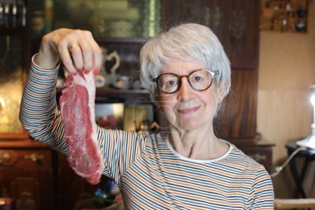 Foto de Close-up portrait of mature woman with raw beef steak at home - Imagen libre de derechos