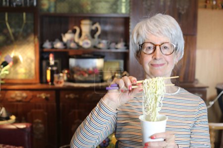 Foto de Close-up portrait of mature woman eating cup noodles at home - Imagen libre de derechos