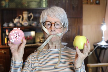 Photo for Close-up portrait of mature woman with doughnut, apple and measuring tape on mouth at home - Royalty Free Image