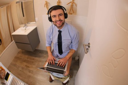Téléchargez les photos : Wide angle shot of young man working with laptop in toilet - en image libre de droit