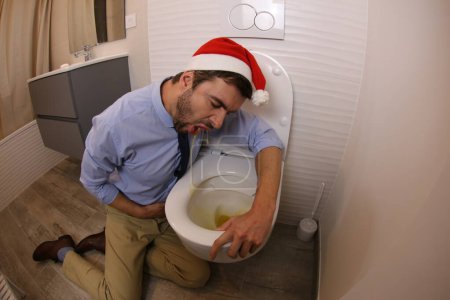 Foto de Wide angle shot of young man vomiting in santa hat in toilet - Imagen libre de derechos