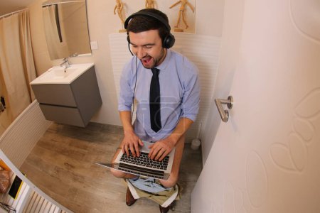 Photo for Wide angle shot of young man working with laptop in toilet - Royalty Free Image