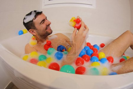 Téléchargez les photos : Plan grand angle de jeune homme heureux avec des canards et des boules en caoutchouc s'amuser dans le bain - en image libre de droit