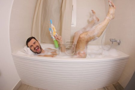Téléchargez les photos : Plan grand angle de beau jeune homme jouant avec pistolet à eau dans le bain - en image libre de droit