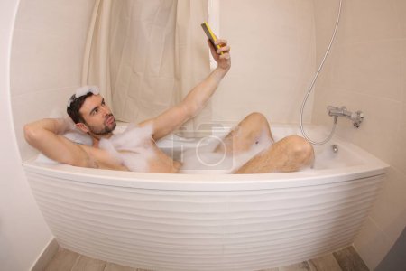 Téléchargez les photos : Plan grand angle de beau jeune homme prenant selfie avec smartphone dans le bain - en image libre de droit