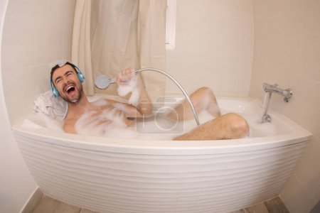 Foto de Amplio ángulo de tiro de joven guapo con auriculares cantando con cabezal de ducha en el baño - Imagen libre de derechos