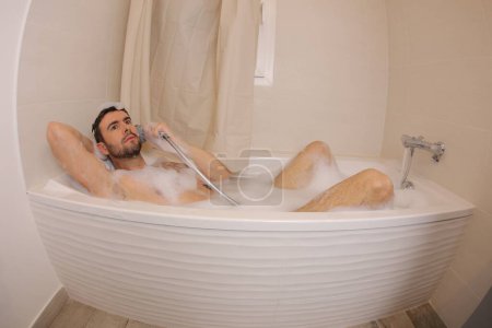 Téléchargez les photos : Plan grand angle de beau jeune homme prétendant parler à travers la pomme de douche comme téléphone dans le bain - en image libre de droit