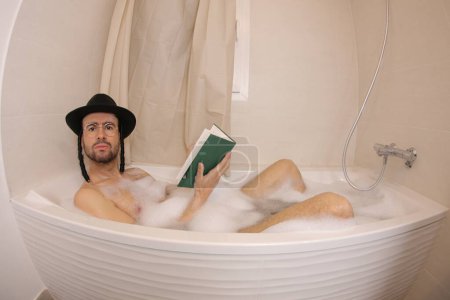 Foto de Wide angle shot of handsome young man in Jewish hat reading tora in bath - Imagen libre de derechos