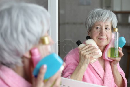 Téléchargez les photos : Portrait en gros plan de femme mature avec des bouteilles de soins de la peau devant le miroir dans la salle de bain - en image libre de droit