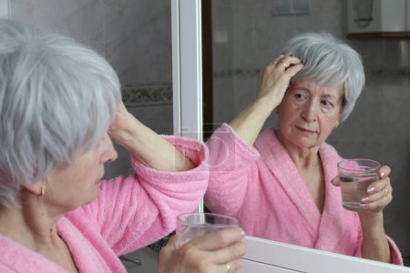 Téléchargez les photos : Portrait en gros plan de femme mature avec des maux de tête tenant verre d'eau devant le miroir dans la salle de bain - en image libre de droit