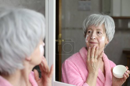 Photo for Close-up portrait of mature woman applying facial cream in front of mirror in bathroom - Royalty Free Image