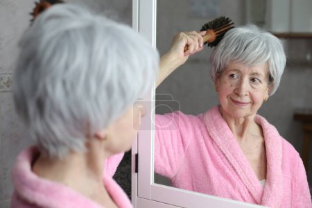 Téléchargez les photos : Portrait en gros plan de femme mature brossant les cheveux devant le miroir dans la salle de bain - en image libre de droit