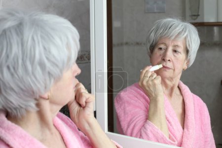 Téléchargez les photos : Portrait en gros plan de femme mature appliquant baume à lèvres devant le miroir dans la salle de bain - en image libre de droit