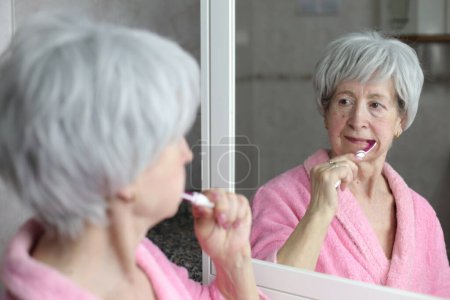 Photo for Close-up portrait of mature woman brushing teeth in front of mirror in bathroom - Royalty Free Image