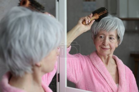 Photo for Close-up portrait of mature woman brushing hair in front of mirror in bathroom - Royalty Free Image