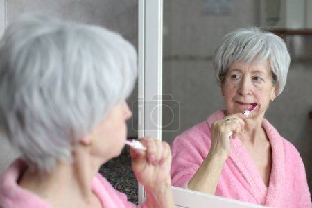 Téléchargez les photos : Portrait en gros plan de femme mature brossant des dents devant le miroir dans la salle de bain - en image libre de droit