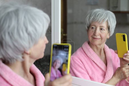 Photo for Close-up portrait of mature woman with smartphone in front of mirror in bathroom - Royalty Free Image