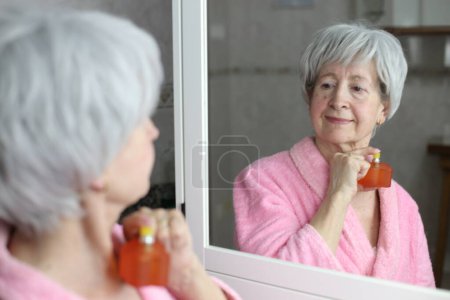Téléchargez les photos : Portrait en gros plan de femme mature appliquant du parfum devant le miroir dans la salle de bain - en image libre de droit