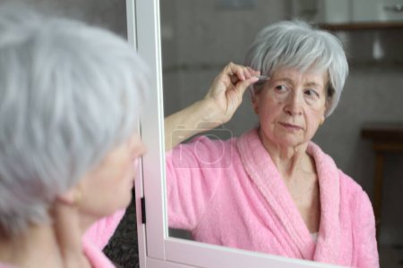 Photo for Close-up portrait of mature woman pulling out facial hair in front of mirror in bathroom - Royalty Free Image