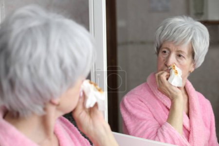 Foto de Retrato de cerca de la mujer madura con la nariz sangrante delante del espejo en el baño - Imagen libre de derechos