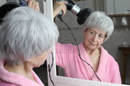 Téléchargez les photos : Portrait en gros plan de femme mature avec sèche-cheveux devant le miroir dans la salle de bain - en image libre de droit