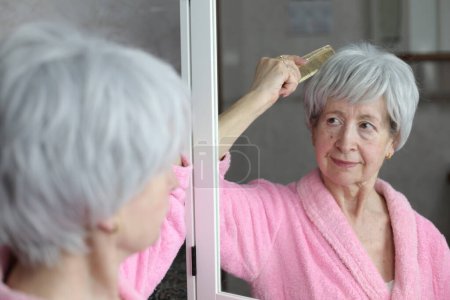 Téléchargez les photos : Portrait en gros plan de femme mature brossant les cheveux devant le miroir dans la salle de bain - en image libre de droit