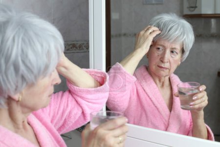 Foto de Primer plano retrato de mujer madura con dolor de cabeza sosteniendo vaso de agua frente al espejo en el baño - Imagen libre de derechos
