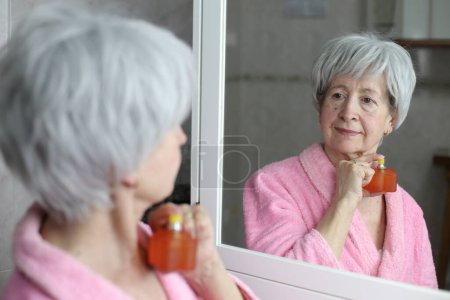 Foto de Primer plano retrato de la mujer madura aplicando perfume delante del espejo en el baño - Imagen libre de derechos