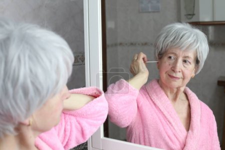 Photo for Close-up portrait of mature woman cleaning ears with cotton sticks in front of mirror in bathroom - Royalty Free Image
