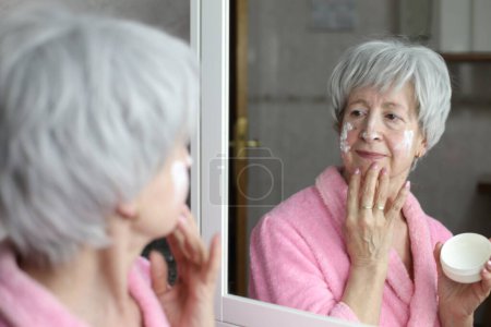 Photo for Close-up portrait of mature woman applying facial cream in front of mirror in bathroom - Royalty Free Image
