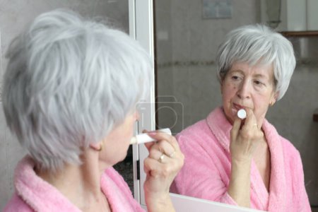 Foto de Primer plano retrato de la mujer madura aplicando bálsamo labial delante del espejo en el baño - Imagen libre de derechos