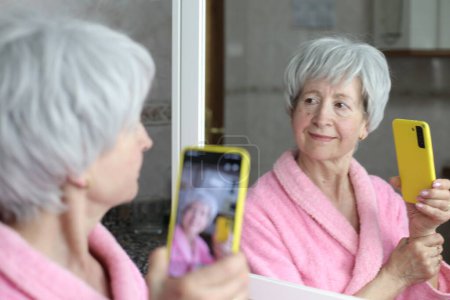 Téléchargez les photos : Portrait en gros plan de femme mature avec smartphone devant le miroir dans la salle de bain - en image libre de droit